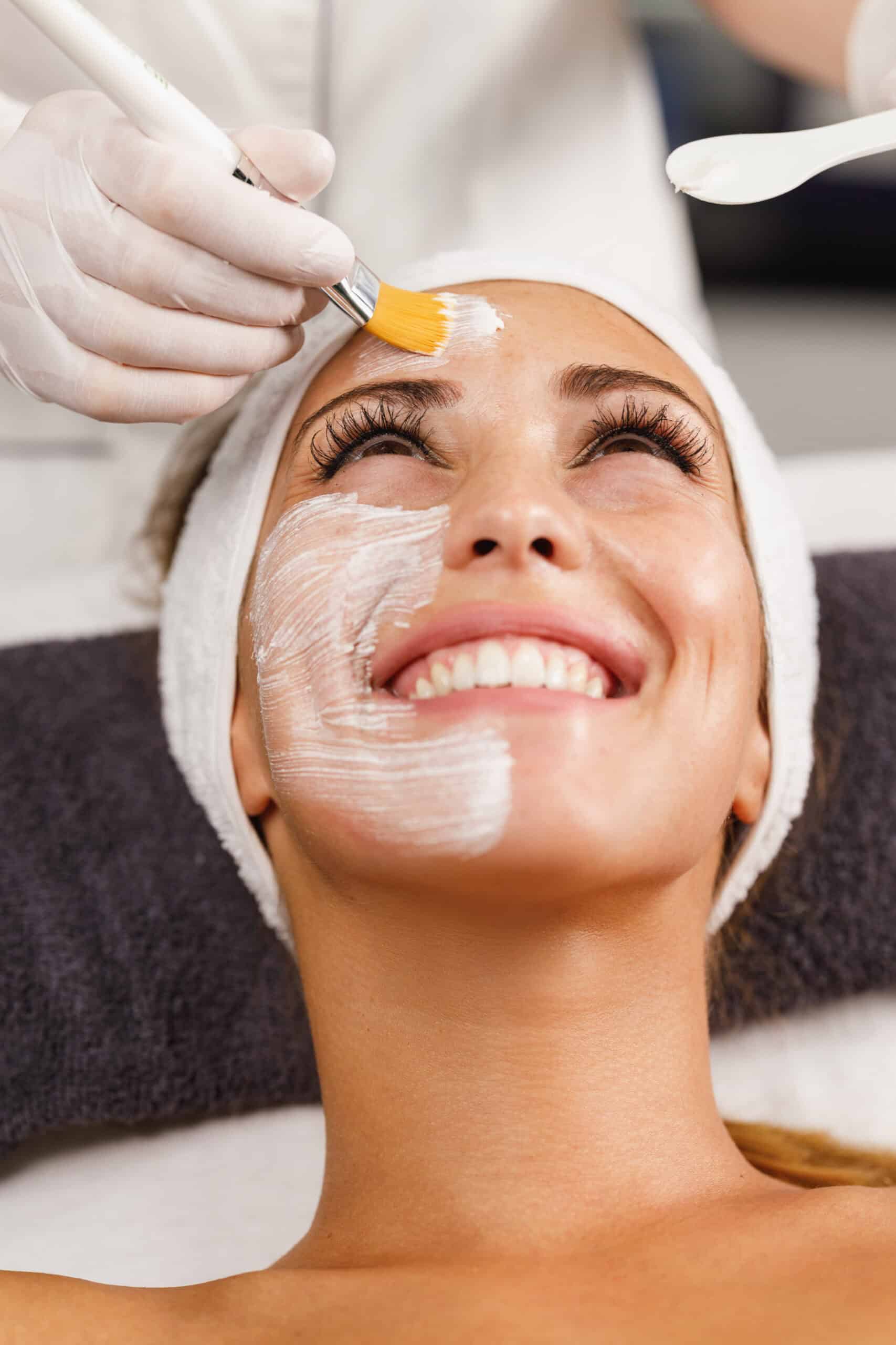 smiling woman looking up as the esthetician brushes a facial cream on her skin