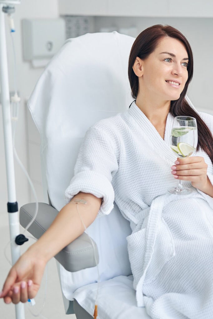 dark haired happy women holding a glass of water while receiving IV therapy
